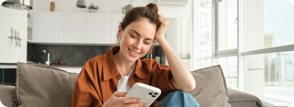 Smiling woman sitting on a couch holding a smartphone in a bright living room.