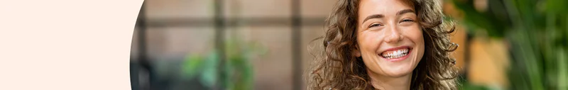 Woman with curly hair smiling in an indoor environment with greenery in the background.