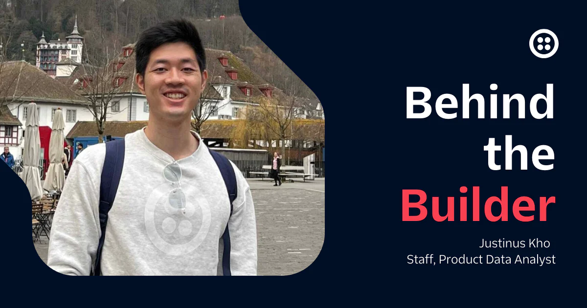 Justinus Kho smiling, wearing a grey sweater and white shirt, with a background of buildings and mountains.