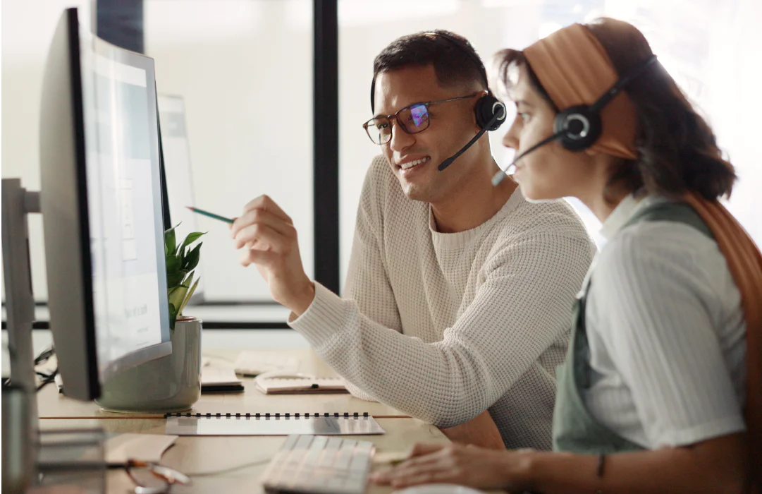 Two customer support representatives wearing headsets collaborating at a desk with a computer.