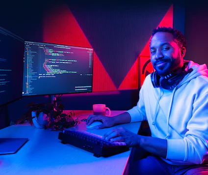 A man wearing headphones working on a dual-monitor computer setup with code displayed on the screens.