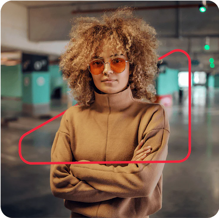 Woman with curly hair and glasses wearing a brown sweater, standing confidently in a parking garage.