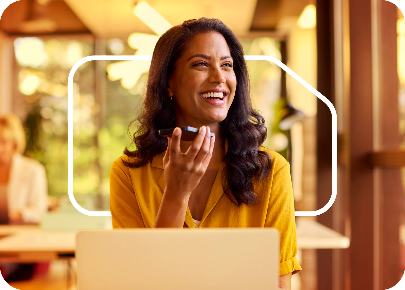 Woman in yellow shirt recording voice message on phone while smiling in a modern office.