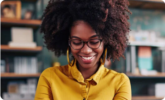 Woman with curly hair, glasses, and yellow shirt smiling in a home office setting.