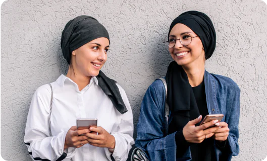 Two women wearing headscarves, smiling and holding smartphones while standing against a textured wall.