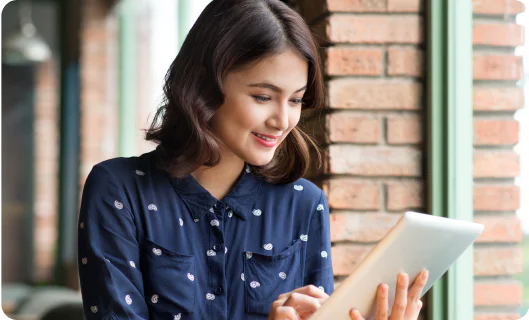 Young woman in a navy shirt smiles while using a tablet by a brick wall with green window frames.