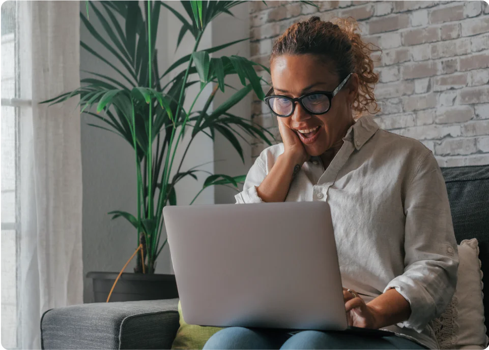A woman with glasses using a laptop and smiling in a cozy living room with a potted plant.
