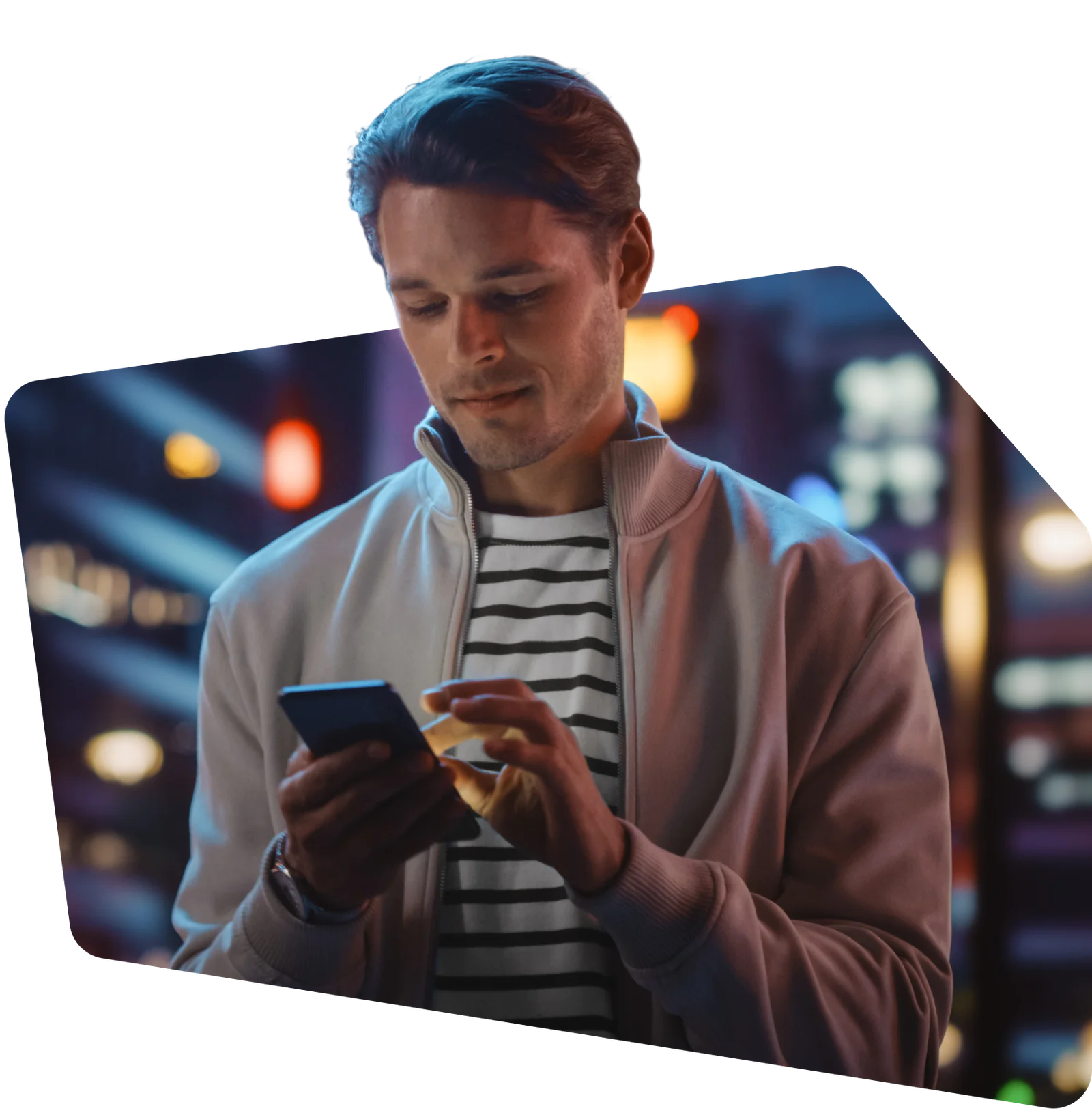 A man in a striped shirt and jacket uses a smartphone in an illuminated urban setting at night.