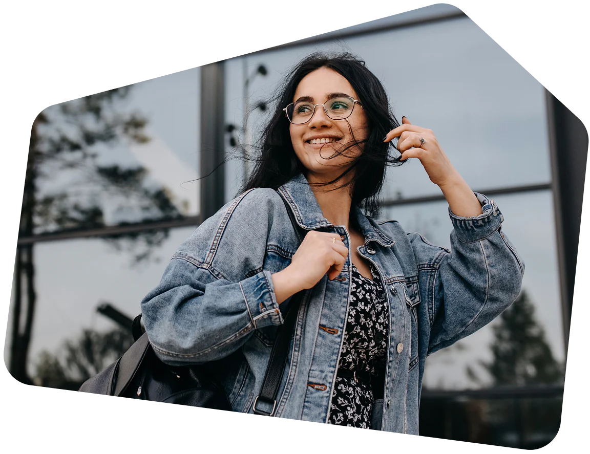Woman with glasses smiling while wearing a denim jacket and carrying a black bag outdoors.