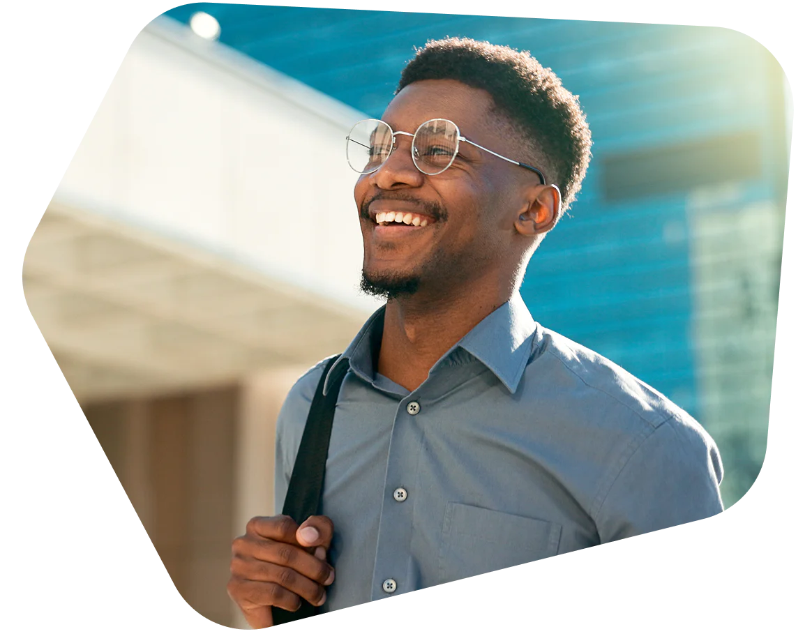 A man with glasses smiling outside, wearing a buttoned shirt and carrying a shoulder bag.