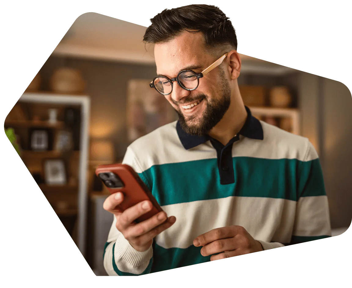 A bearded man with glasses smiling at his phone in a cozy room.