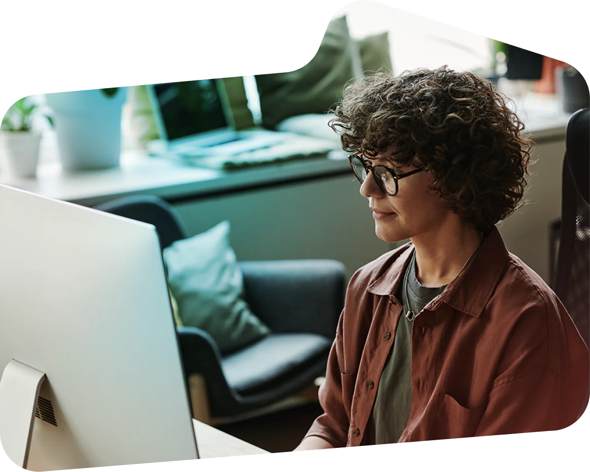 Woman with curly hair and glasses working on a computer in a modern office environment.