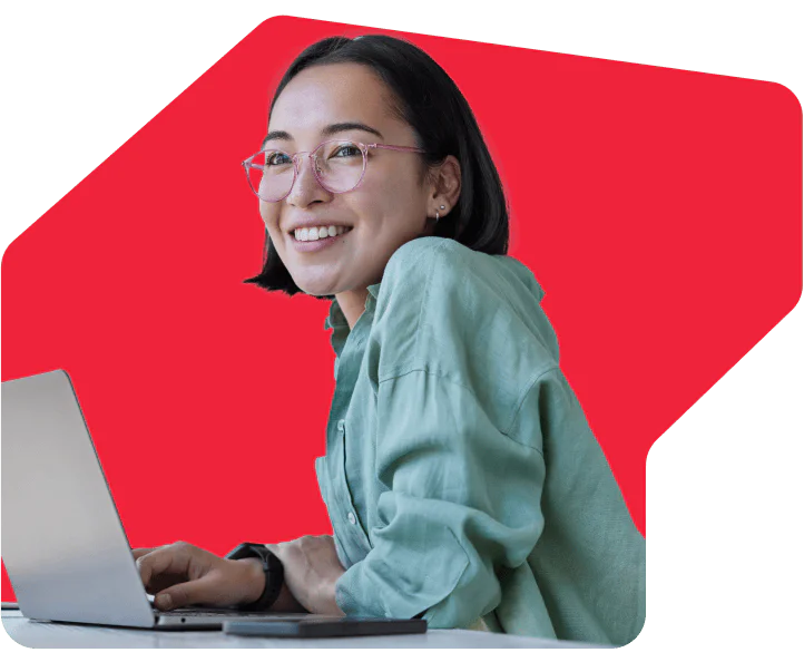 Woman with glasses smiling while using a laptop, with a red background behind her.