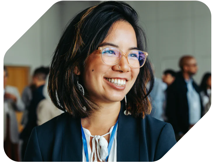 Woman with glasses and a nose ring smiling at an event, with people in the background.