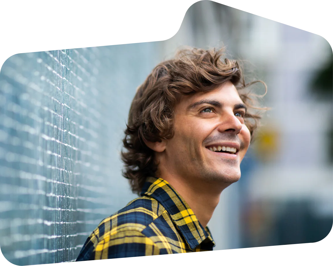 Man with curly hair in a yellow plaid shirt smiling next to a brick wall