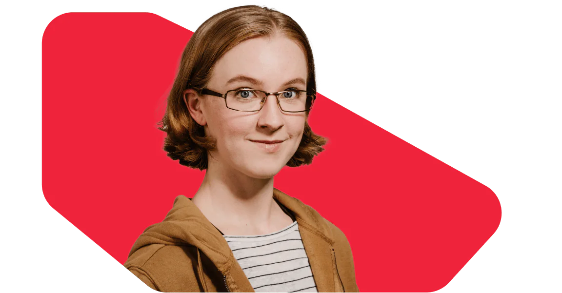 Young woman with glasses and a brown jacket standing in front of a red backdrop, smiling.