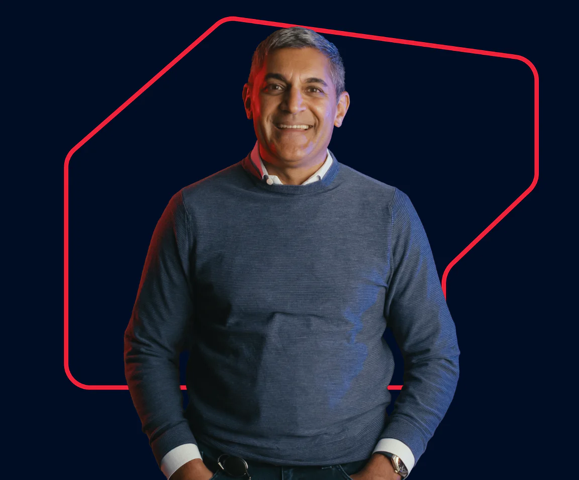 Smiling man in a blue sweater standing with hands in pockets against a dark background with red geometric lines.