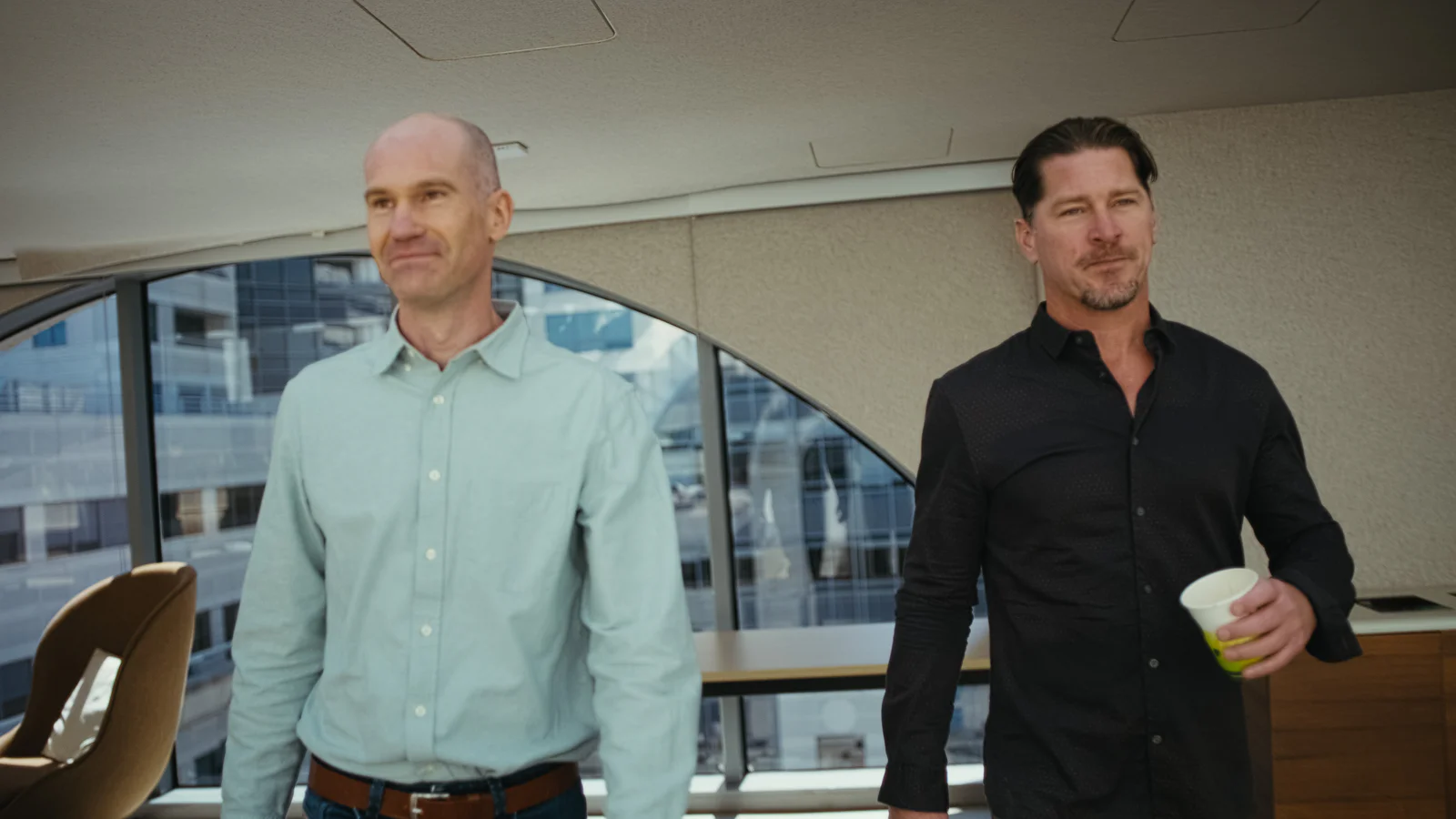 Two men, one in a light blue shirt and the other in a black shirt, walking inside an office with large windows.