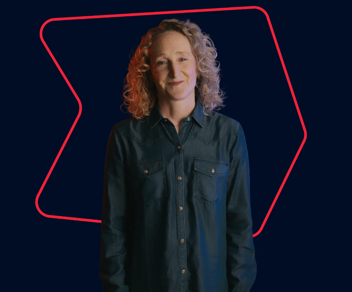 Smiling woman with curly hair wearing a denim shirt stands against a dark background with a neon outline.