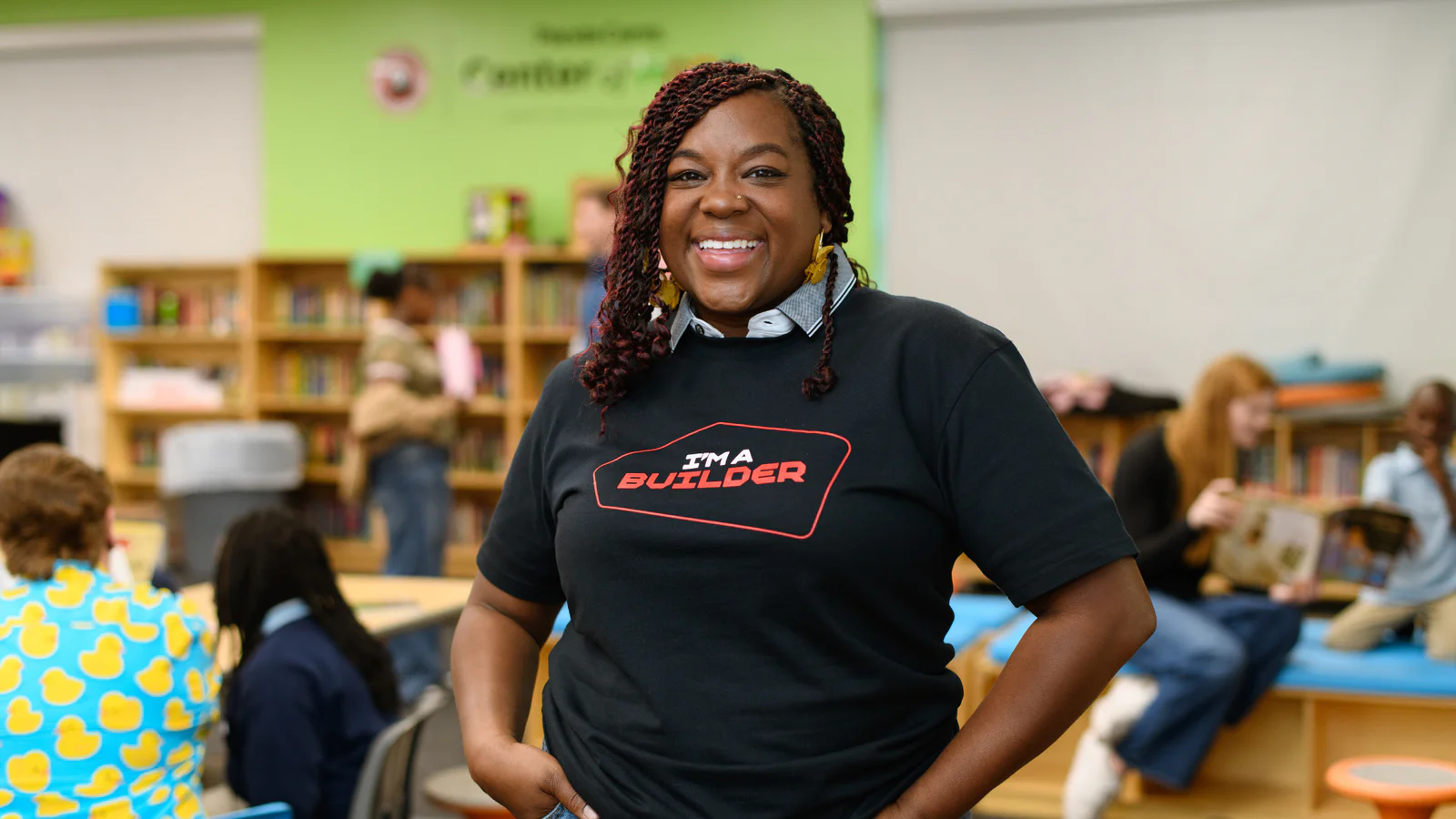 Smiling woman wearing I'm a Builder shirt in a classroom with students in the background
