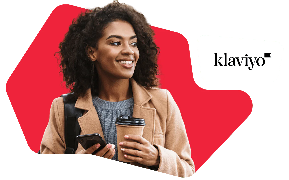 Woman with curly hair holding a coffee cup and smartphone, wearing a tan coat, smiling.